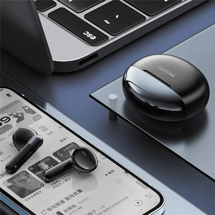 Black wireless earbuds with charging case on a desk next to a smartphone and laptop.